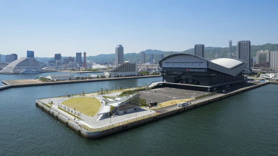 神戸の港湾エリアを上空から捉えた写真。GION ARENA KOBEを中心に、整備された公園や、特徴的な建物が並ぶ市街地、そして海が広がる美しい景観が写し出されています。晴れた日の開放的な風景です。