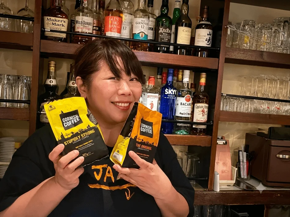 バーカウンターを背景に、笑顔の女性が「CORNISH COFFEE」のコーヒー豆の袋を2つ持っています。棚には様々な種類の酒瓶が並べられています。