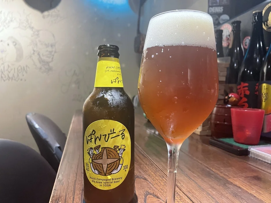 高知カンパーニュブルワリーの「ぱんびーる」のボトルと、泡立ったビールが注がれたグラスが木製のテーブルに置かれた写真です。居酒屋のような雰囲気の中で、クラフトビールを楽しんでいる様子が伝わります。