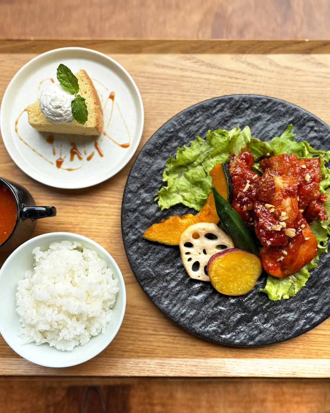 木製のトレーに盛り付けられた定食とデザートの画像です。メインは鶏肉料理と彩り豊かな野菜（かぼちゃ、レンコン、さつまいも、オクラ）で、ご飯とスープが添えられています。デザートはホイップクリームとミントが飾られたケーキにキャラメルソースがかかっています。