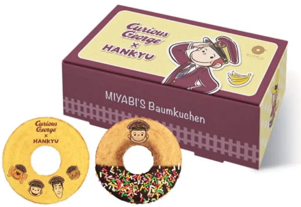 おさるのジョージと阪急電鉄のコラボレーション商品「MIYABI'S Baumkuchen」の箱とバウムクーヘンが写っています。車掌姿のジョージがデザインされ、チョコレートとスプリンクルで飾られたものも含まれます。