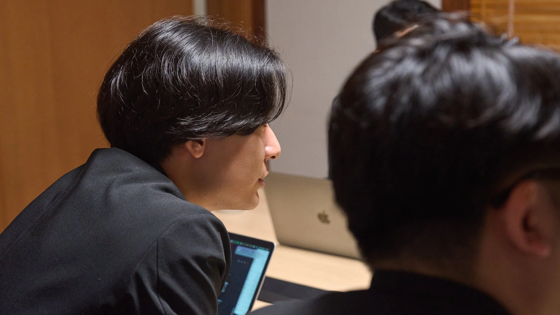 会議室のような場所で、黒いジャケットを着た男性がノートパソコンに向かっている横顔