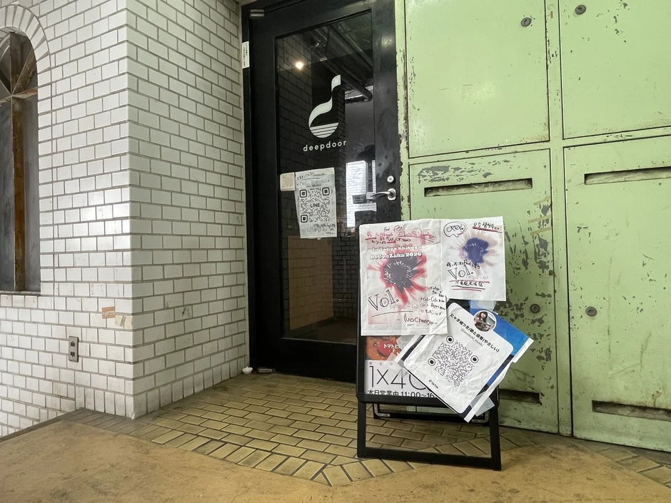 白いレンガ壁の建物にある「deepdoor」という店の入り口。手書きのチラシが貼られた看板があり、QRコードやイベント「Vol.1」、営業時間が表示されている。隣には古い緑色のロッカーが並ぶ。