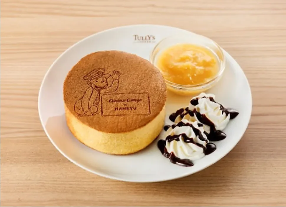 白い皿に盛り付けられた、おさるのジョージの焼き印が特徴的な厚焼きパンケーキ。フルーツソースとホイップクリーム、チョコレートソースが添えられており、タリーズと阪急のコラボレーションスイーツである。