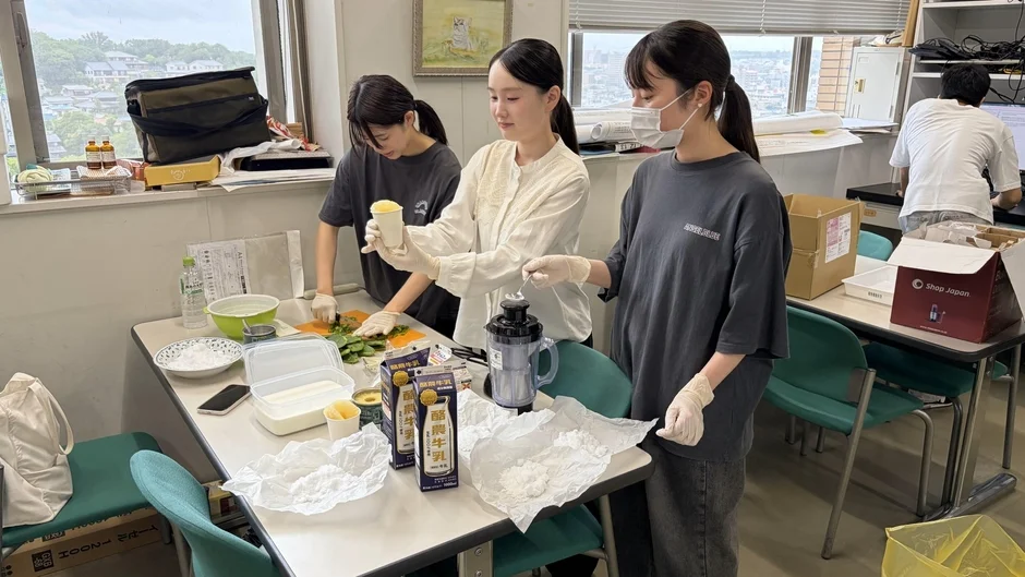 室内で3人の女性が手袋を着用し、テーブル上で牛乳や野菜を使ってドリンクを準備している。ブレンダーも使用されており、共同で調理作業を行っている様子が伺える。