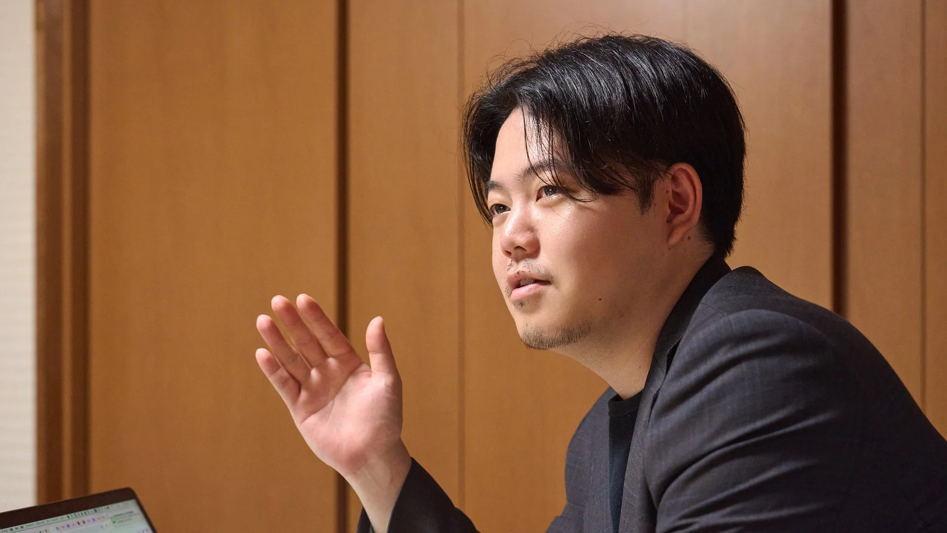 黒いスーツを着た若いアジア人男性が、室内で身振り手振りを交えながら話している様子