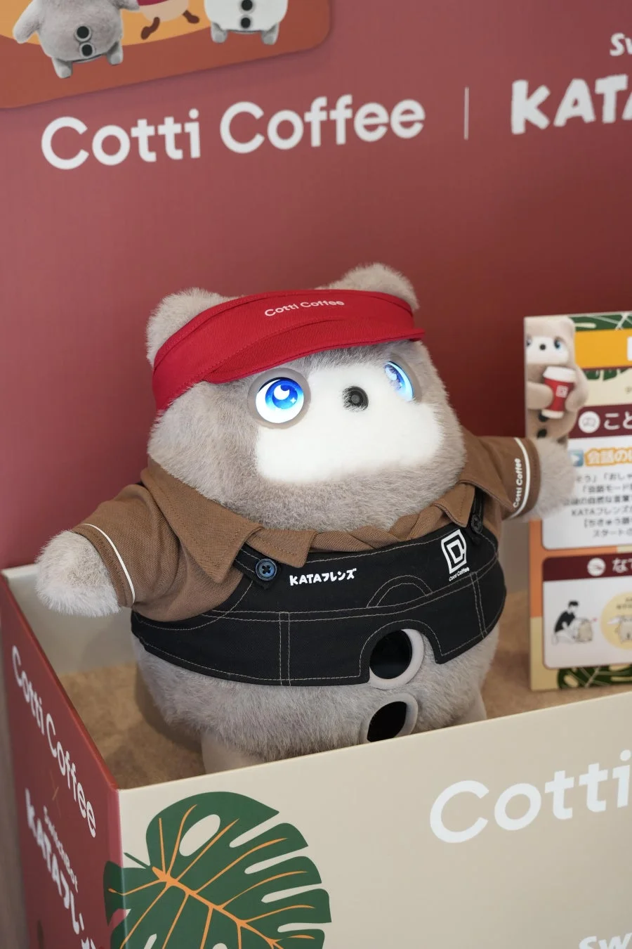 Cotti Coffeeの制服を着たKATAフレンズのロボットが、Cotti Coffeeのロゴが入った箱に展示されています。