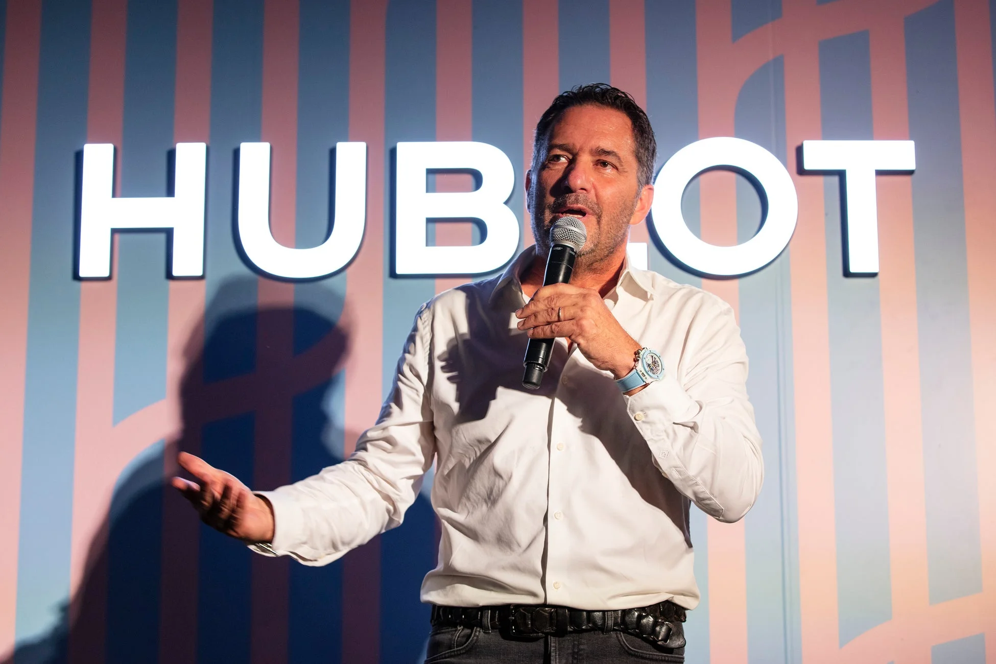 HUBLOTのロゴを背景に、白いシャツを着た男性がマイクを持ってスピーチ