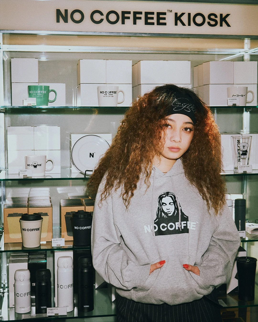 NO COFFEE KIOSKのディスプレイの前で、NO COFFEEのロゴ入りパーカーを着た巻き髪の女性が立っています。棚にはマグカップやタンブラーなどのブランドグッズが並んでいます。