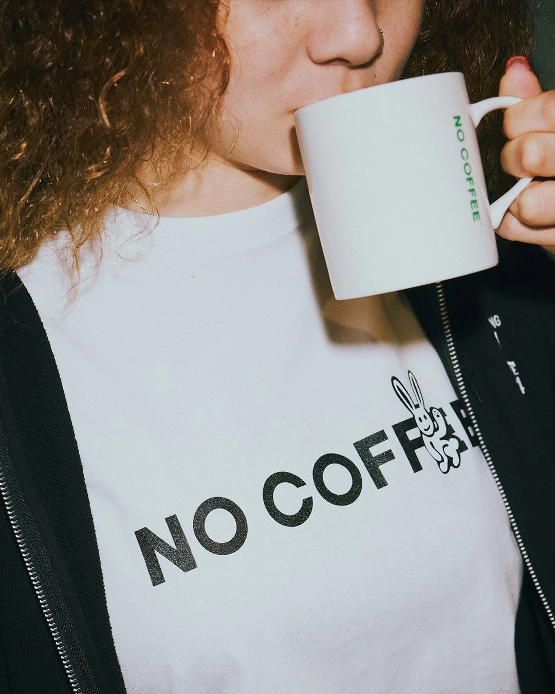 カーリーヘアの人物が白いマグカップで飲み物を飲んでいます。着用している白いTシャツには「NO COFFEE」の文字とウサギのイラストが、マグカップにも「NO COFFEE」のロゴが見えます。