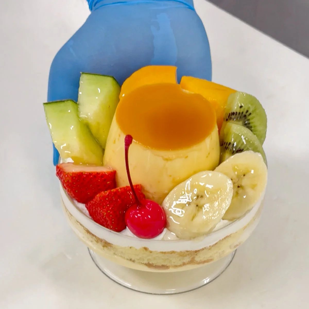 青い手袋をはめた手が、カラメルソースがかかったプリンと色とりどりのフルーツをたっぷり使った華やかなカップデザートを持っている