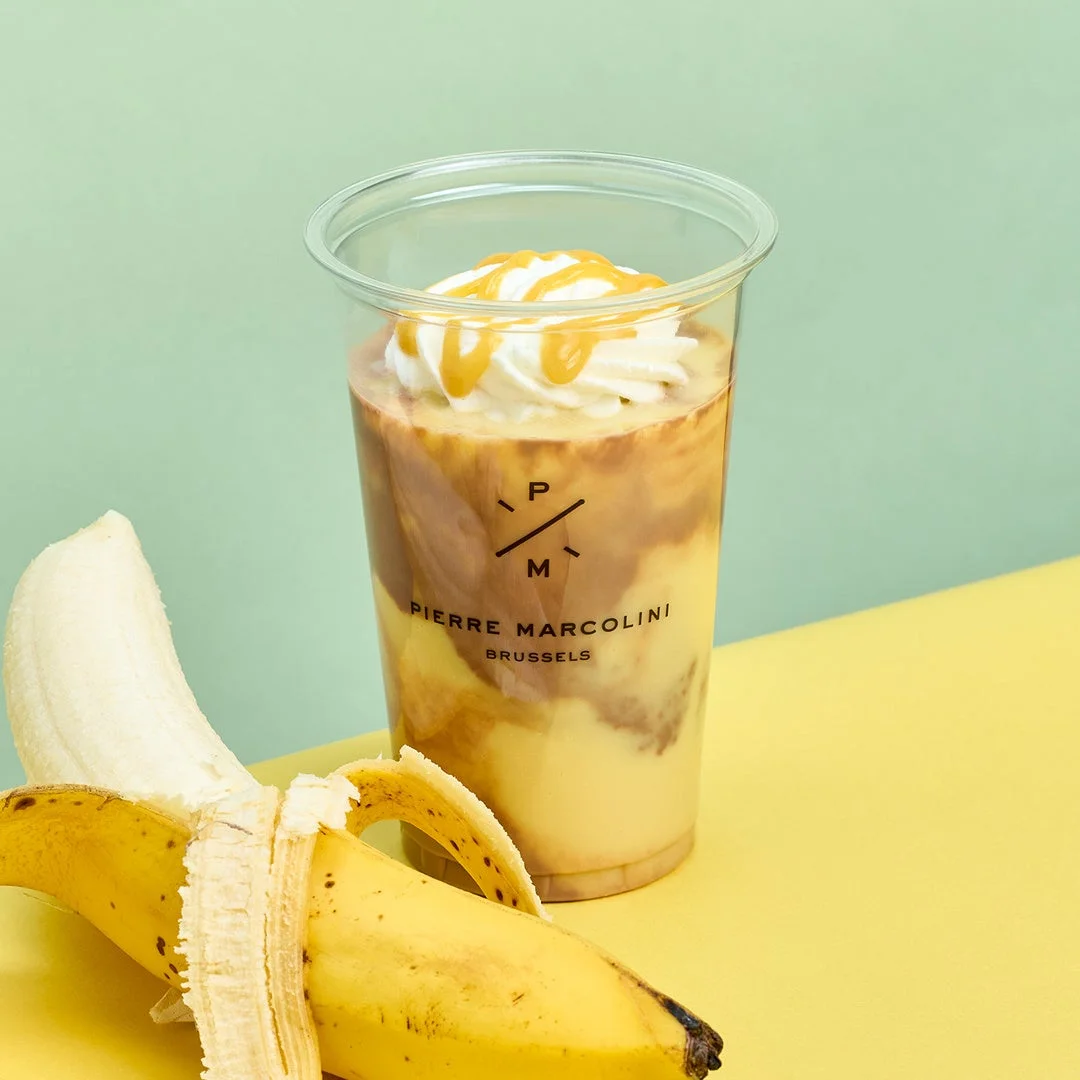 ピエールマルコリーニのロゴ入りカップに入った、バナナとチョコレートのような層が美しいドリンクです。ホイップクリームとキャラメルソースがトッピングされており、隣には皮をむいたバナナが置かれています。