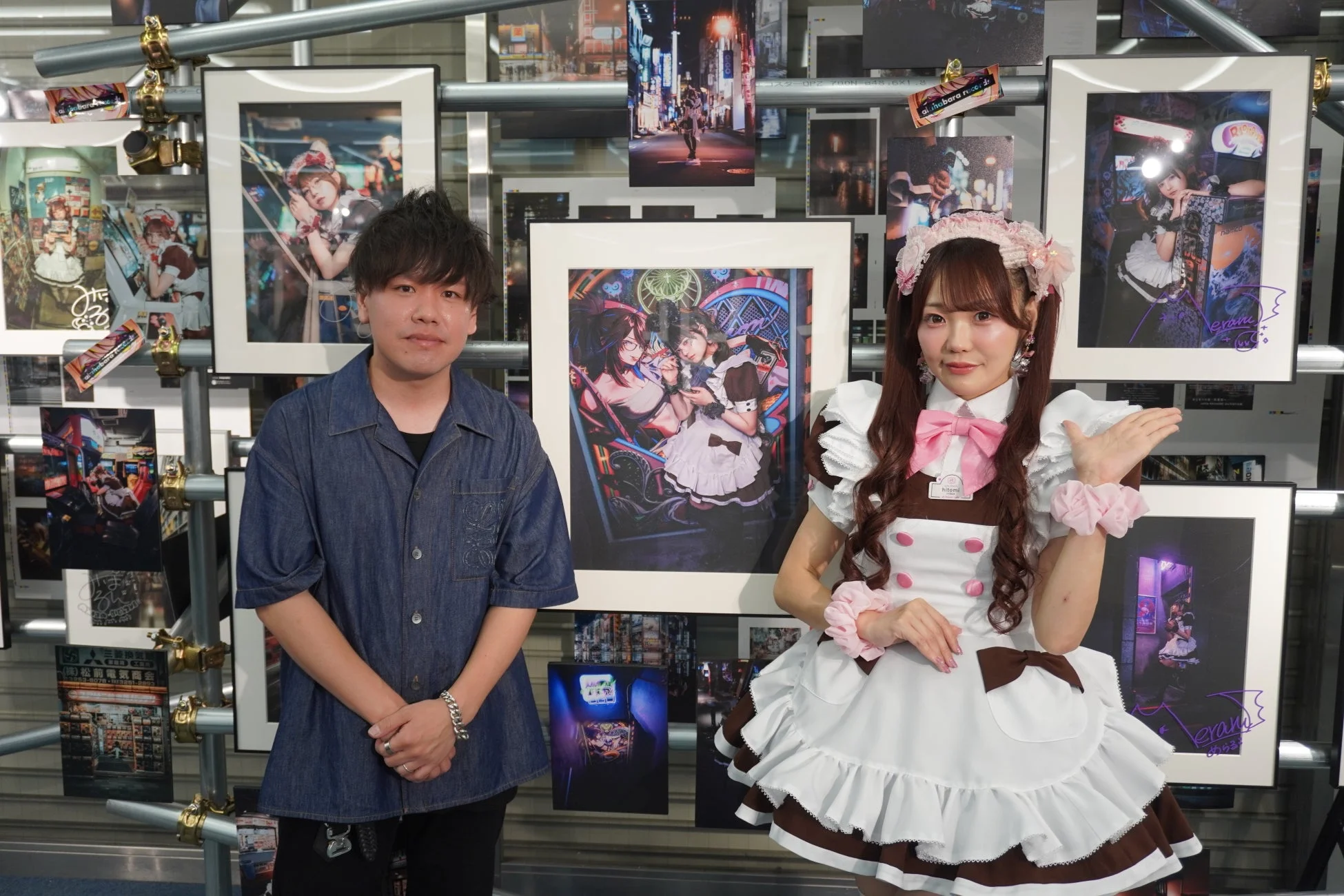 メイド服の女性と男性が、メイドや秋葉原をテーマにした写真やイラストが飾られた展示会の壁の前でポーズをとっている。