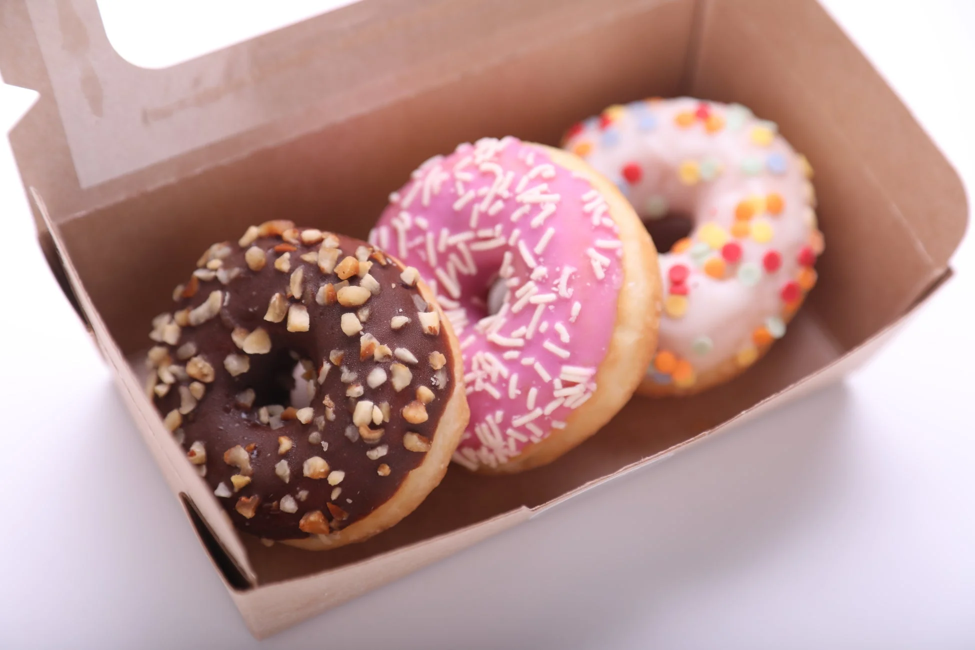 段ボールの箱に、チョコレート、ピンク、白いアイシングが施された3つのカラフルなドーナツが入っています。それぞれナッツやスプリンクルで飾られ、美味しそうなスイーツのセットです。
