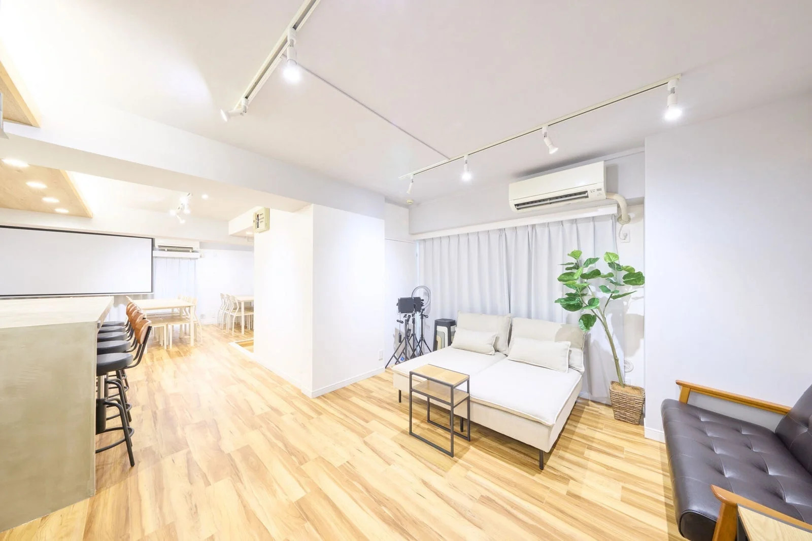 白い壁と木目調のフローリングが特徴の、明るく広々としたリビングダイニング空間。バーカウンター、プロジェクタースクリーン、ソファ、ダイニングセットが備えられ、多目的に利用できるモダンな内装の部屋