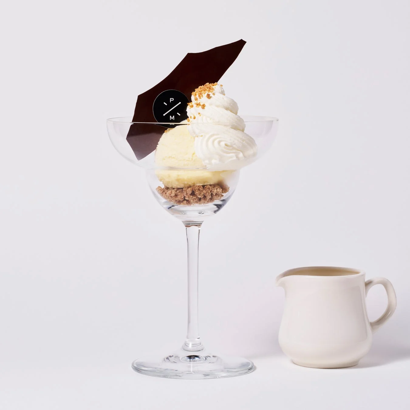 白い背景に置かれた、脚付きグラスに入ったアイスクリームとホイップクリームのデザート。横には小さなミルクピッチャーが添えられています。