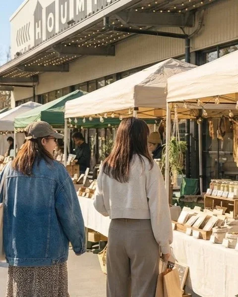 晴れた日に屋外マーケットを訪れる二人の女性が写っています。多くの屋台が並び、人々が買い物を楽しんでいる様子が伺えます。