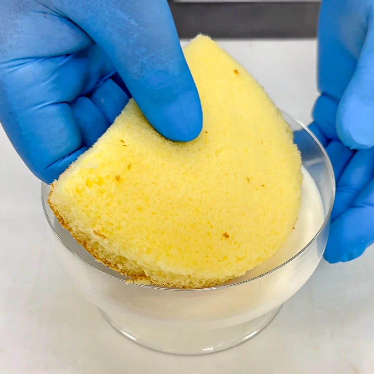 青い手袋をはめた手が、ふわふわのスポンジケーキを白い液体が入った透明なグラスに浸している