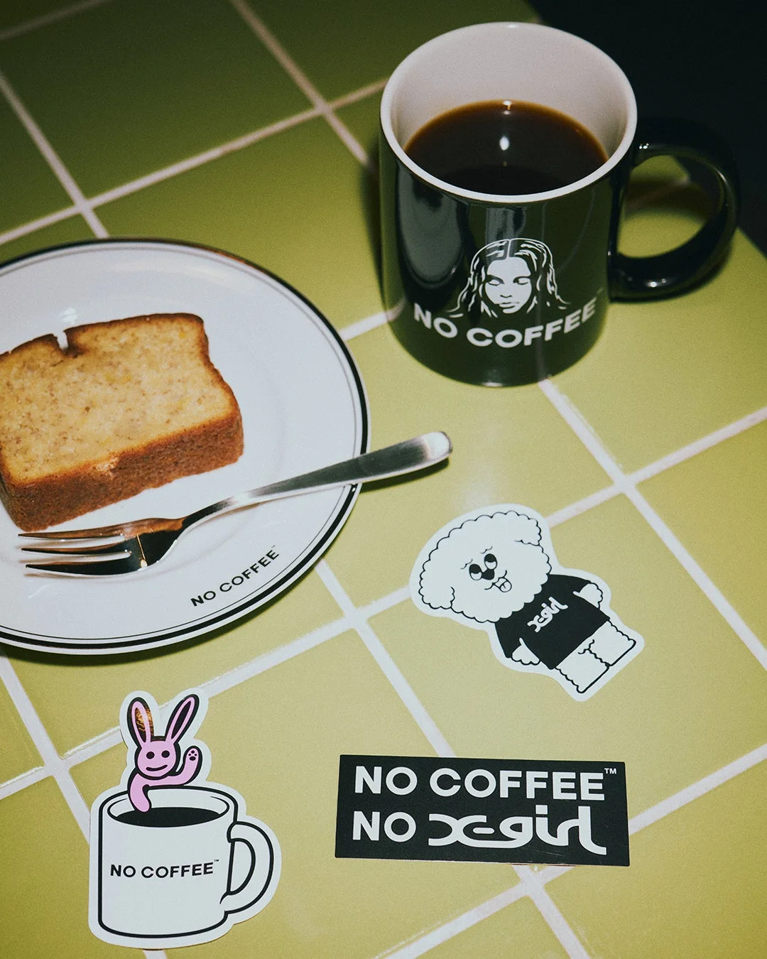 緑のタイル張りのテーブルに、コーヒーマグとバナナブレッドが置かれ、NO COFFEEとxgirlのコラボレーションステッカーが散りばめられたカフェブレイクの様子です。