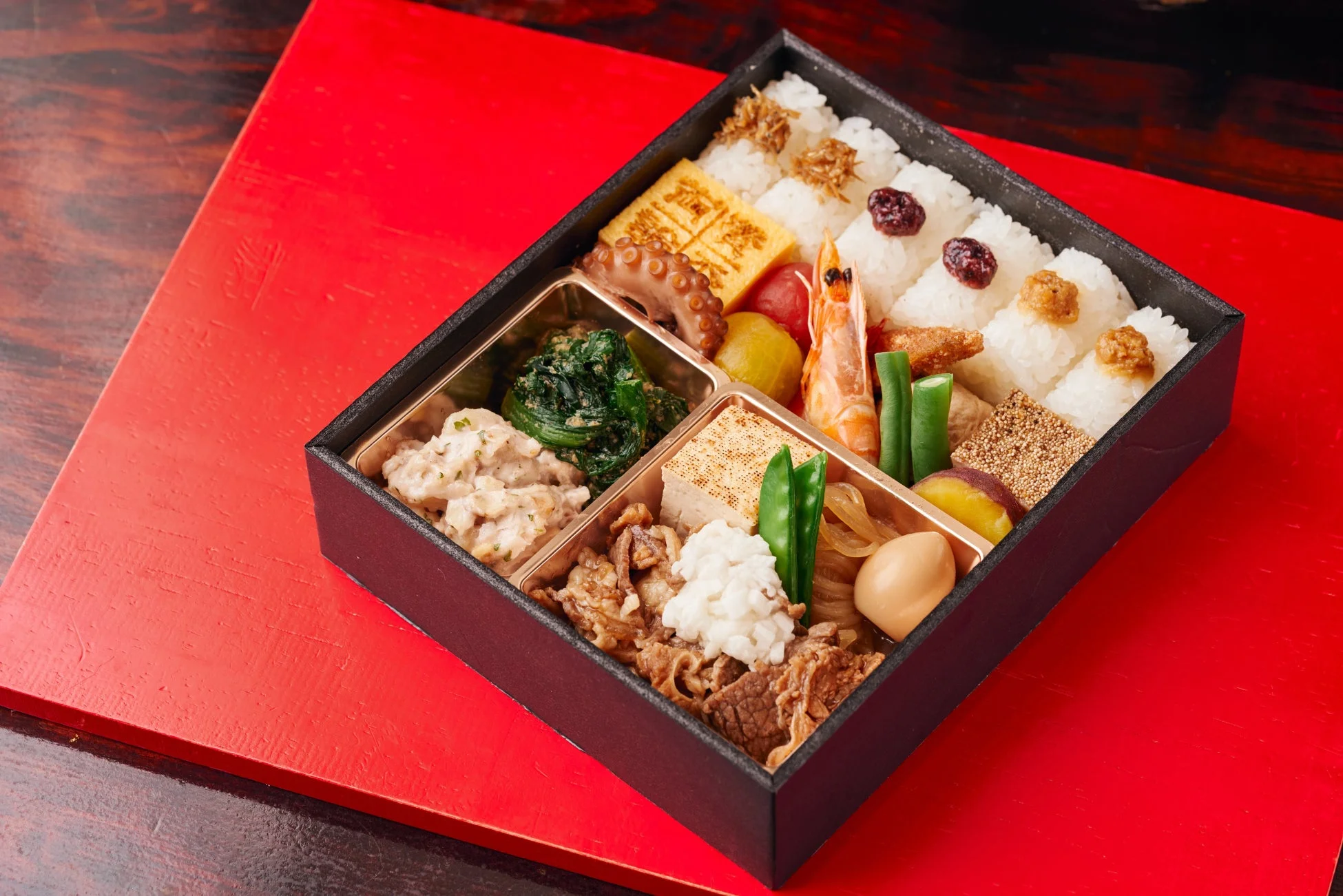 黒い弁当箱に、おにぎり、玉子焼き、海老、タコ、牛肉など、彩り豊かな和食のおかず