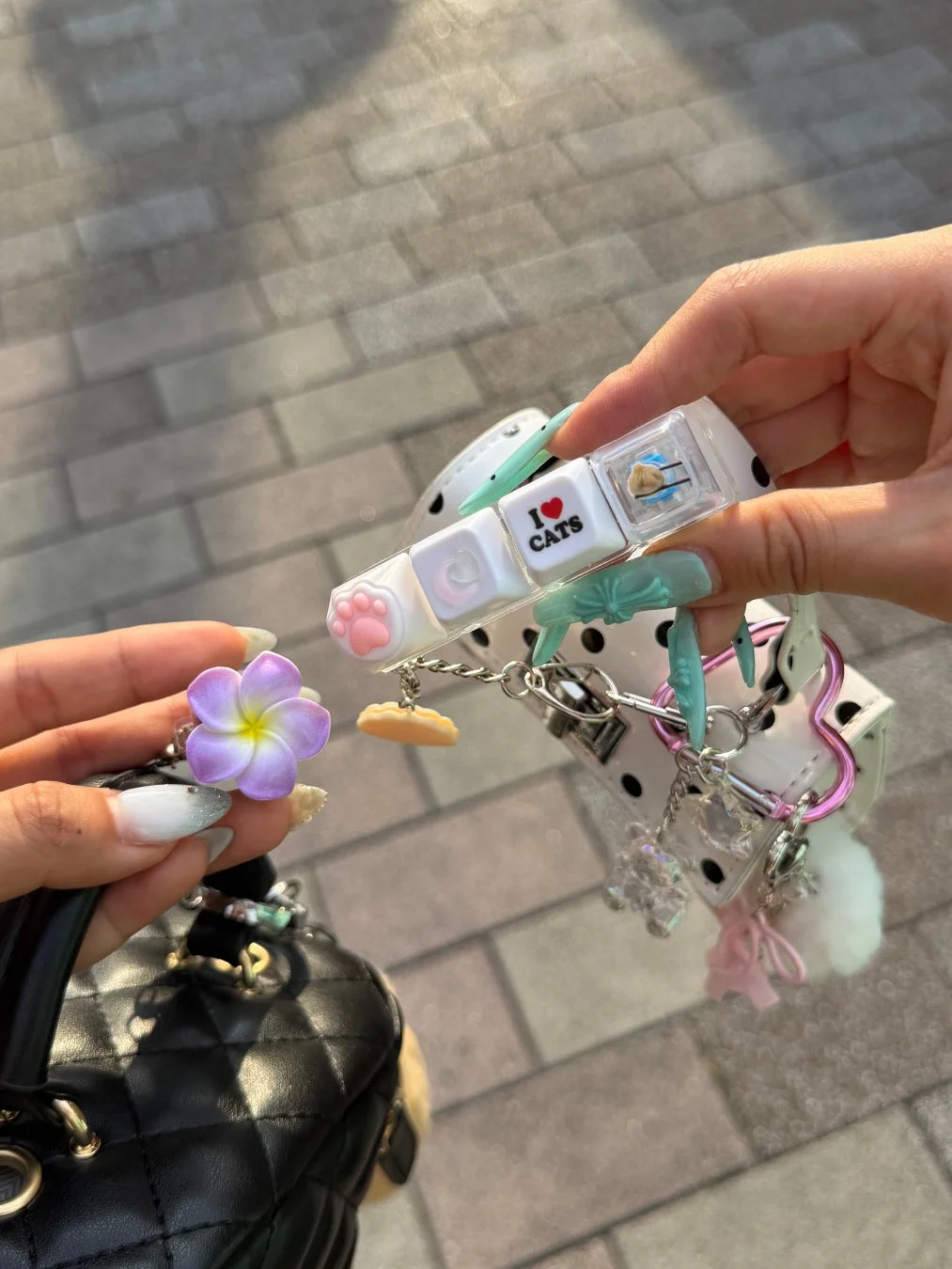 2つの手が、猫の肉球や「I LOVE CATS」の文字が入ったキーボード風のチャーム付きアクセサリーと、キルティングバッグに付いた花のチャームを持っている様子。