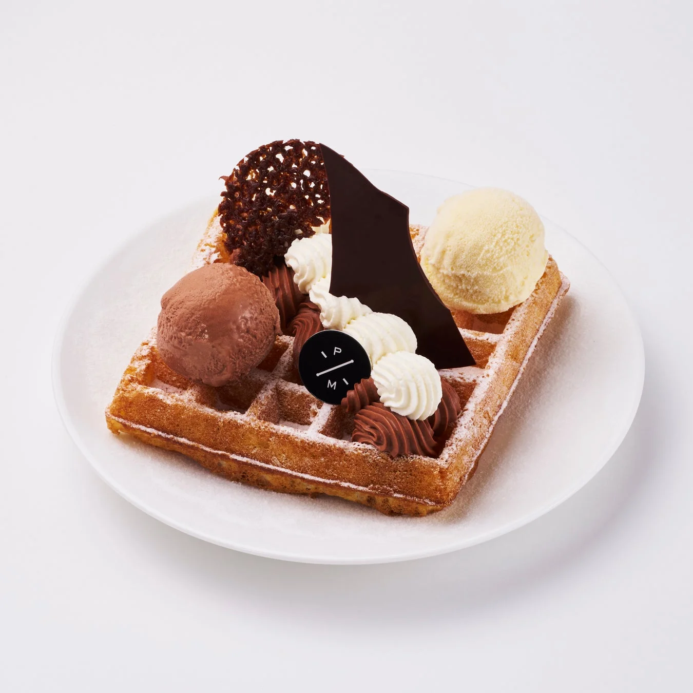 白い皿に盛られたワッフルデザート。チョコレートとバニラのアイスクリーム、2色のホイップクリーム、チョコレートの飾り付けが施されており、粉砂糖がかけられた華やかな一品です。