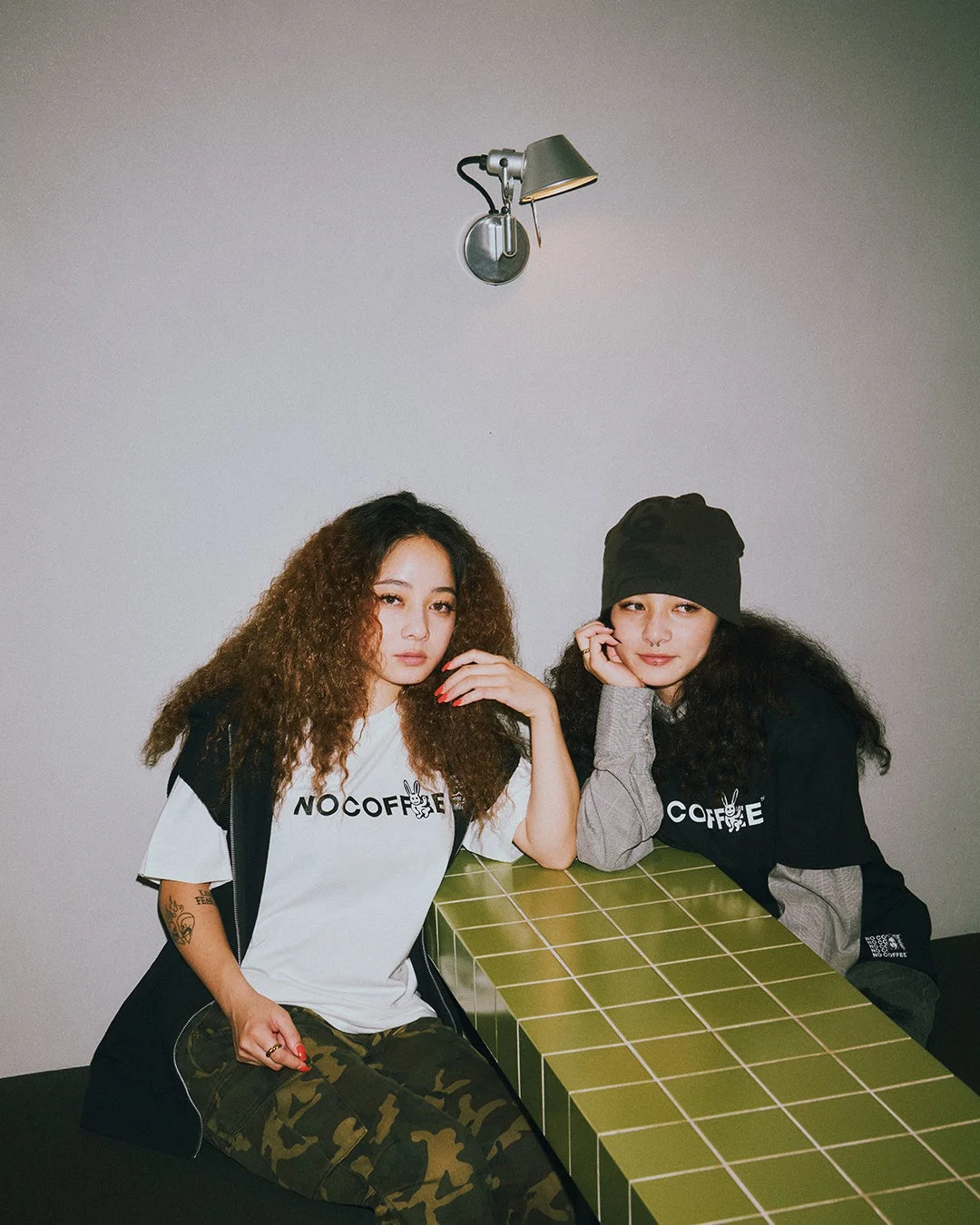 緑色のタイル張りのテーブルに座る、カーリーヘアの若い女性2人のポートレート。カジュアルなストリートウェアファッションで、Tシャツには「NO COFFEE」のロゴが見える。