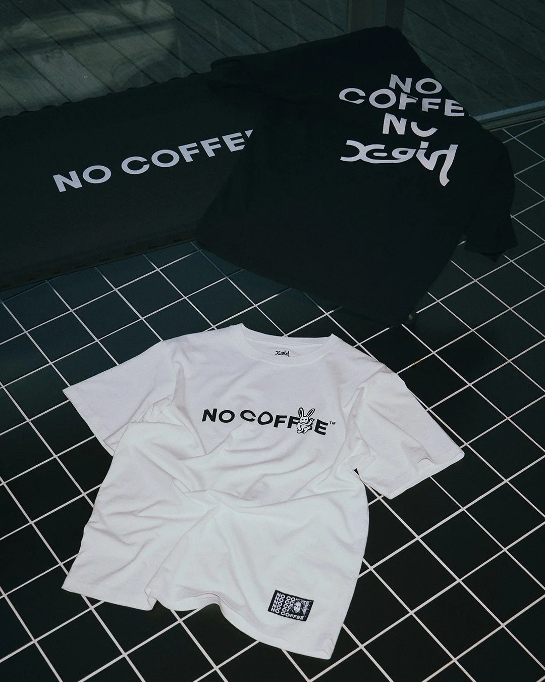 黒いタイル張りの床に、NO COFFEEとX-girlのコラボTシャツが3枚並べられています。白と黒のTシャツにはそれぞれのブランドロゴや、ウサギのキャラクターがデザインされています。