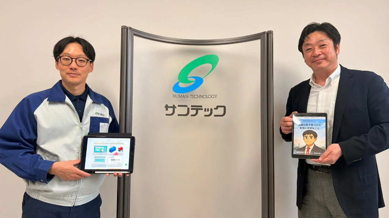 サンテックのロゴを背景に、2人の男性がタブレットを手に立っている。左の男性は作業着、右の男性はスーツ姿で、タブレットには会社の資料やアニメイラストが表示され、ビジネスシーンを連想させる。