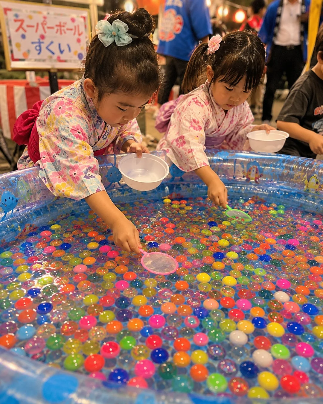 浴衣を着た2人の幼い女の子が、お祭りでスーパーボールすくいを楽しんでいる様子。カラフルなスーパーボールが浮かぶプールから、真剣な表情でボールをすくっている。日本の夏祭りや縁日の賑やかな雰囲気が伝わる一枚。