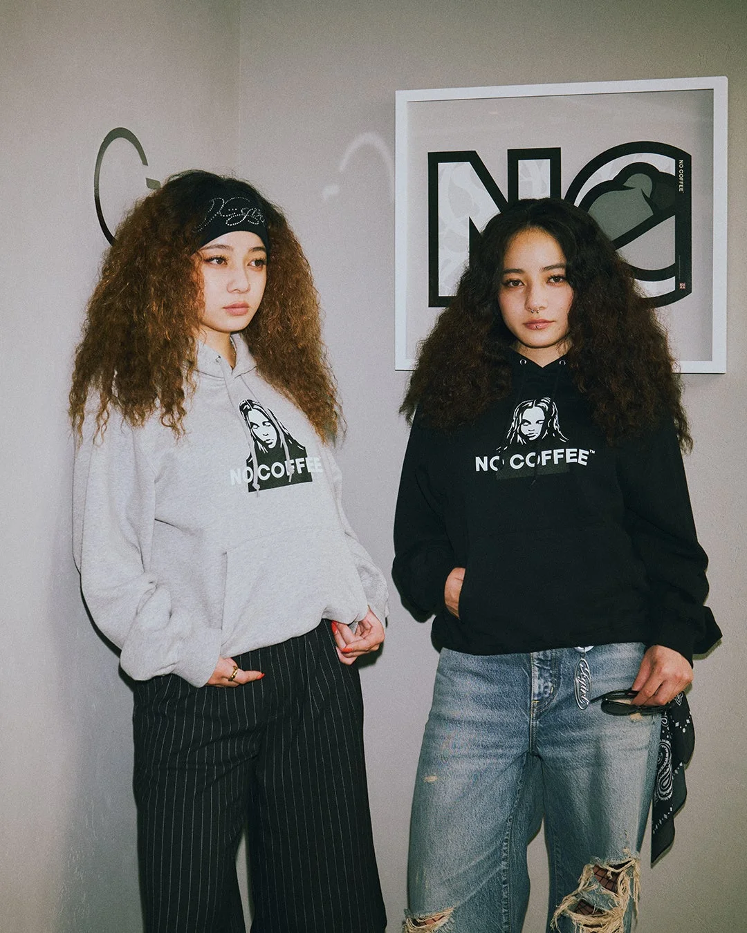 NO COFFEEのロゴ入りパーカーを着たカーリーヘアの女性二人が並ぶポートレート。グレーと黒のパーカーに、ピンストライプパンツとダメージジーンズを合わせたクールなストリートファッションが特徴。