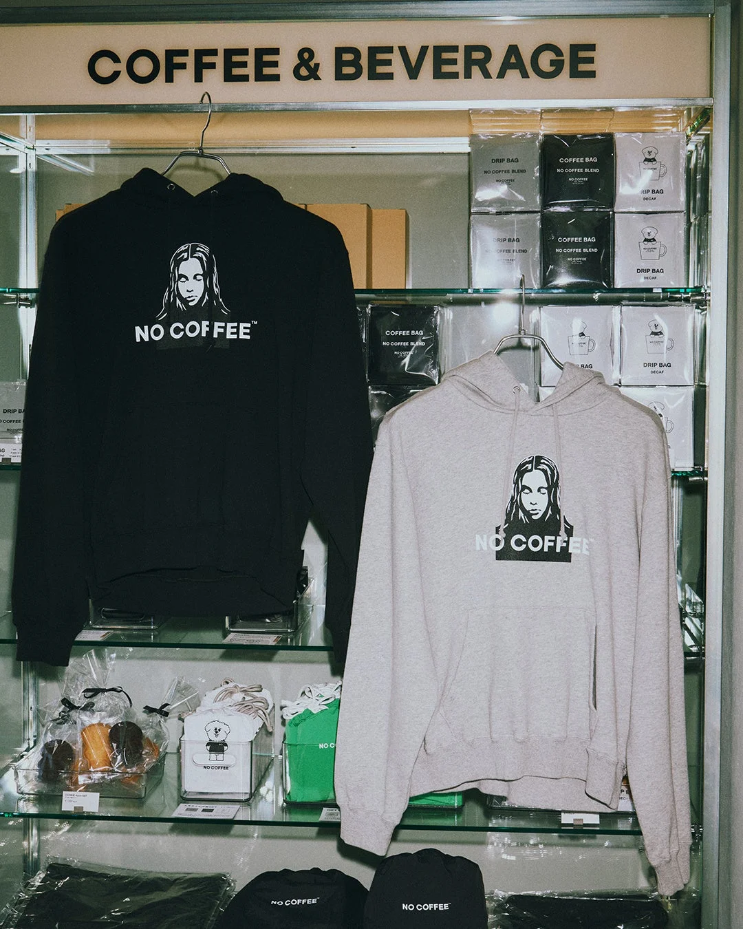 「COFFEE & BEVERAGE」のサインがある棚に、「NO COFFEE」のロゴが入った黒とグレーのパーカー、ドリップバッグコーヒー、その他グッズが陳列されている様子。コーヒー関連のアパレルや商品を販売する店舗の一角です。